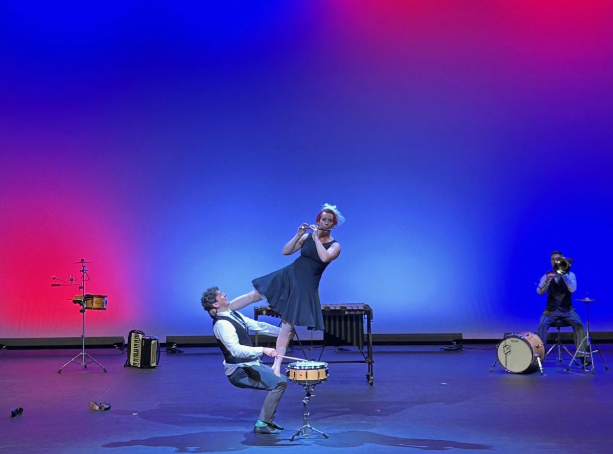 The Fourth Wall’s Hilary Abigana playing the flute while balancing on Greg Jukes who is playing the snare drum with C. Neil Parsons playing the trombone in the background for “Fruit Flies Like a Banana” in The Theatre at Doudna Performing Arts Center