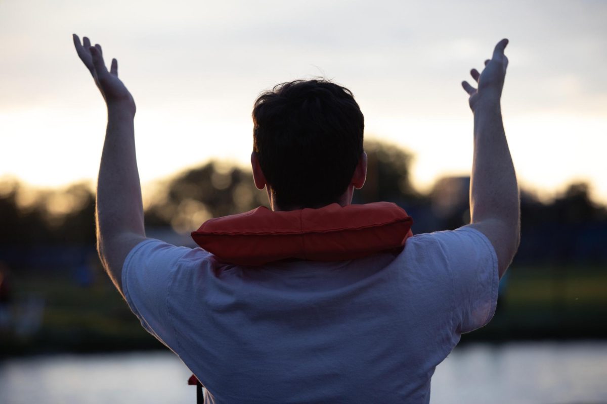 Junior history education major John Petrovich raises his hand to the team on the other side of Campus Pond to tell them to come to the other side, during the neighborhood week Boat race 